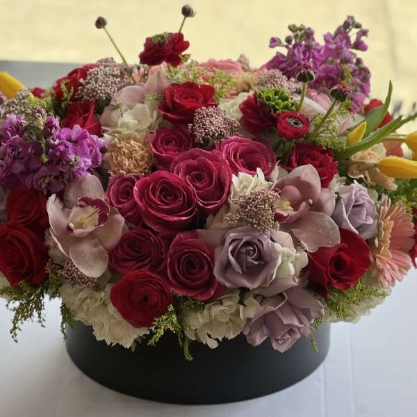 Round arrangement of pink and red roses with purple blooms in a black box