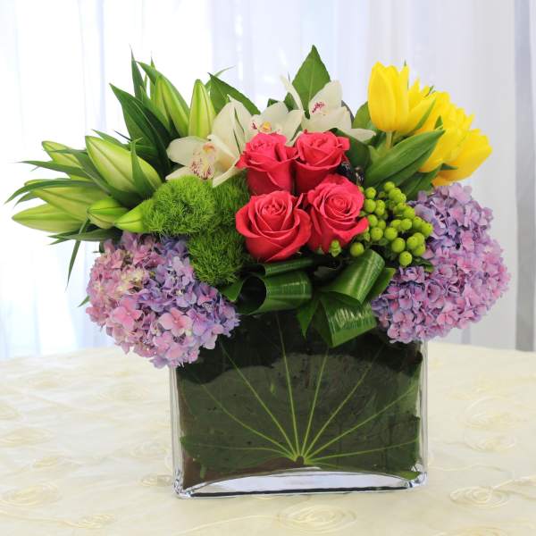 Mixed bouquet in a clear square vase with roses, lilies, hydrangeas, and tulips