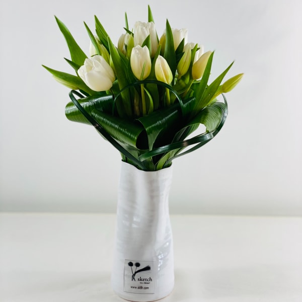 White tulips arranged in a tall white vase with folded green leaves