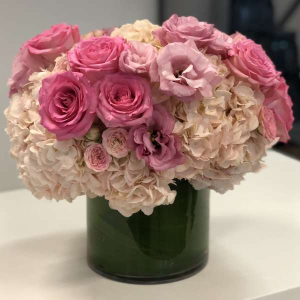 Low round arrangement of pink roses and lisianthus with blush hydrangeas in a glass cylinder vase