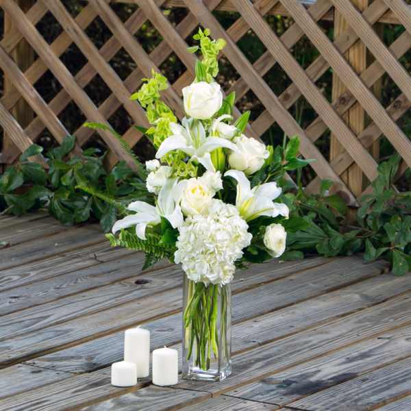 White floral arrangement in a tall glass vase with three candles nearby