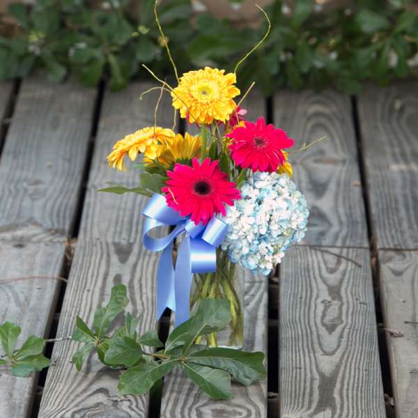 Small bouquet of pink and yellow gerbera daisies with blue hydrangea in a glass vase