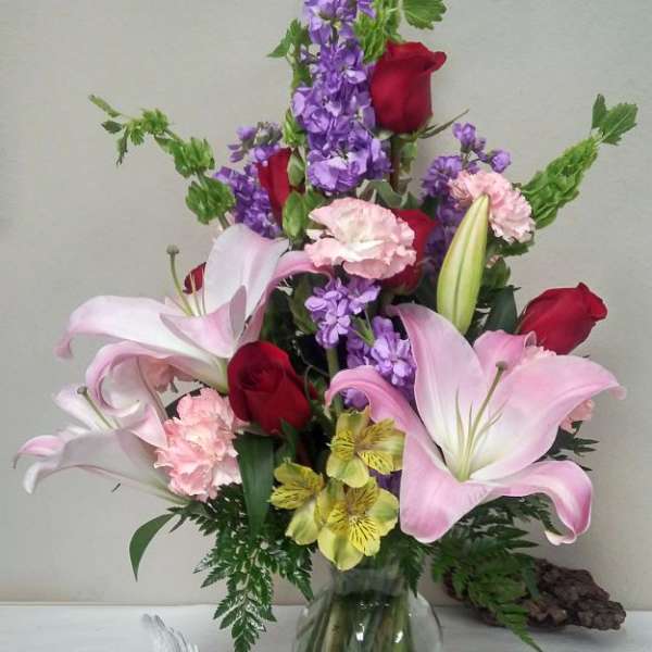 Mixed bouquet of pink lilies, red roses, purple flowers, and carnations in a glass vase