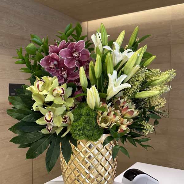 Tall arrangement of orchids, lilies and protea in a textured gold vase on a marble counter