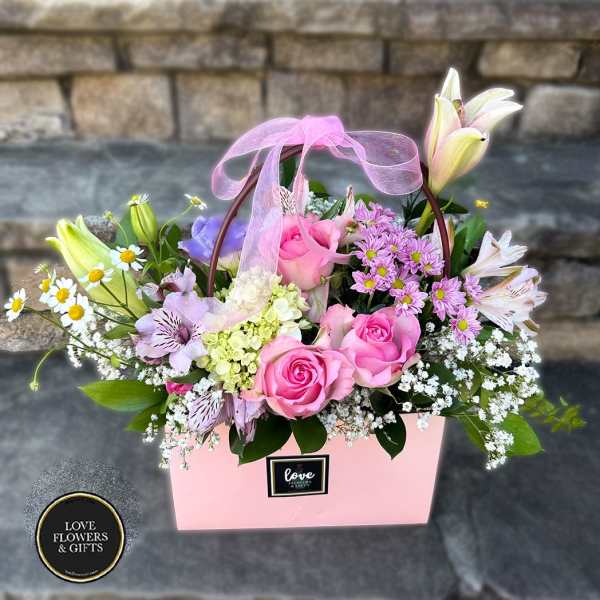 Pink gift bag of mixed flowers including pink roses, lilies, daisies, and filler with a sheer pink bow.