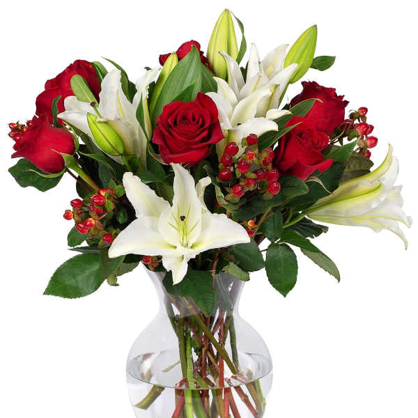 Red roses and white lilies arranged with red berries in a clear glass vase