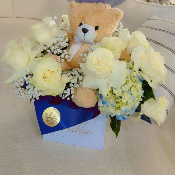 Teddy bear in white roses with a blue welcome balloon