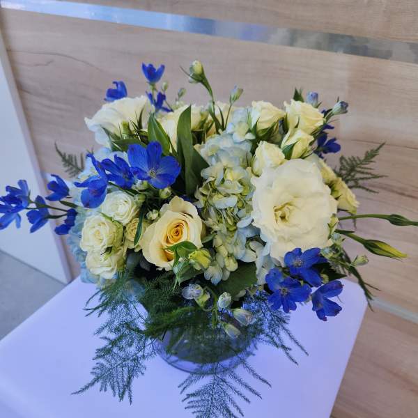 Low round arrangement of white and cream flowers with blue accents in a clear glass vase