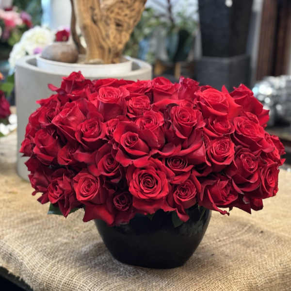 Low arrangement of many red roses densely packed in a glossy black bowl.