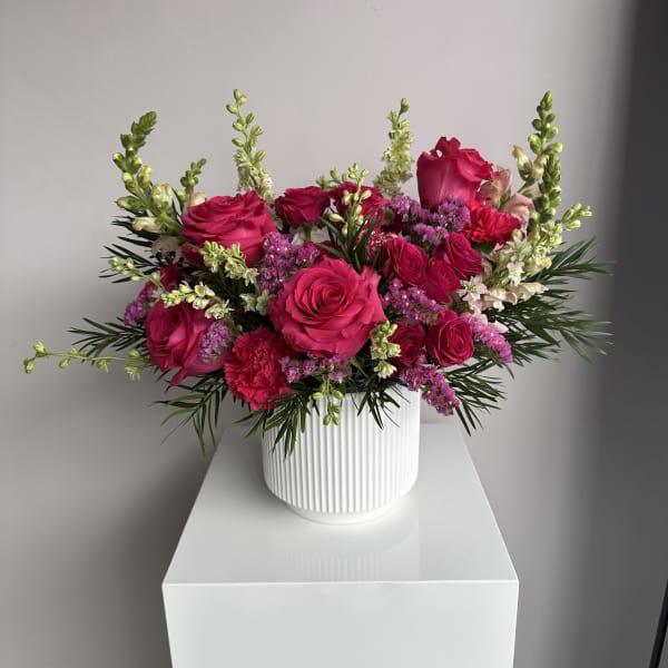 Bright pink rose and carnation arrangement with white snapdragons in a white ribbed ceramic vase.