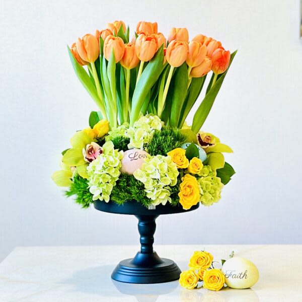Orange tulips in a black pedestal arrangement with yellow roses and green hydrangeas