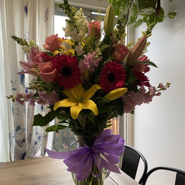 Mixed bouquet of pink, yellow, and white flowers in a glass vase with a purple bow