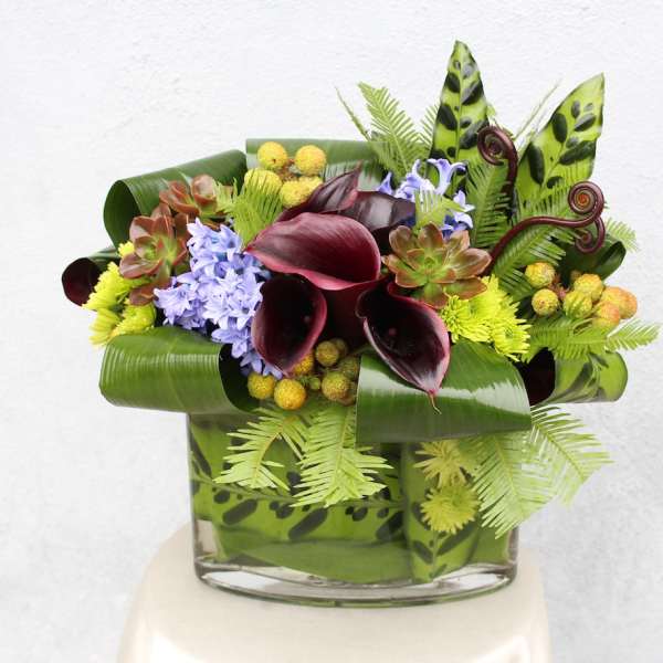Tropical floral arrangement with dark calla lilies and purple blooms in a glass vase