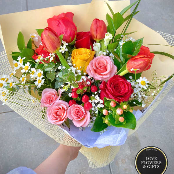 Hand-tied bouquet of mixed roses, tulips, and small white flowers wrapped in pale yellow paper