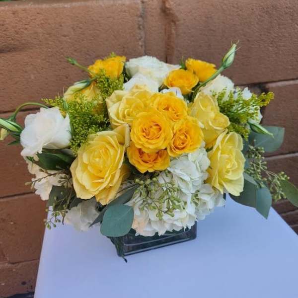 Low arrangement of yellow and white roses with white hydrangeas in a clear glass cube vase