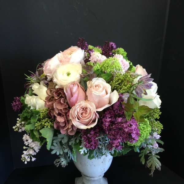 Pastel bouquet of roses, ranunculus, and purple blooms in a white urn vase