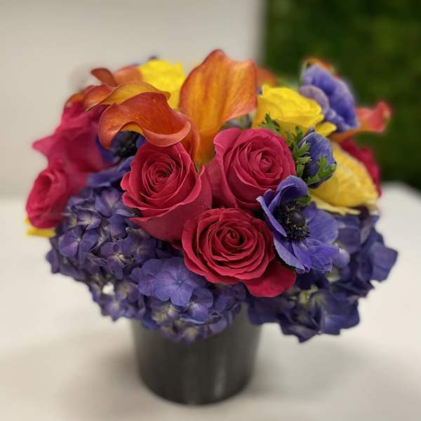 Low round arrangement of hot pink roses, orange calla lilies, yellow roses and purple blooms in a black vase