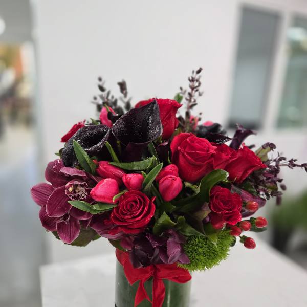Red roses and dark purple flowers arranged in a glass vase with a red ribbon.