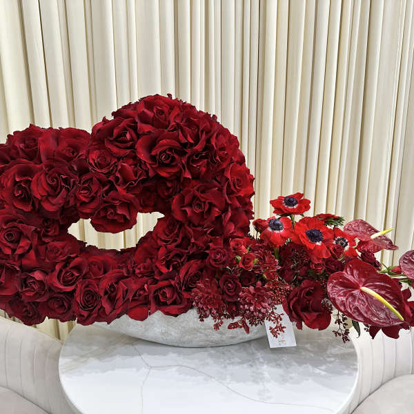 Heart-shaped arrangement of red roses with red blooms and anthuriums in a low white vessel.