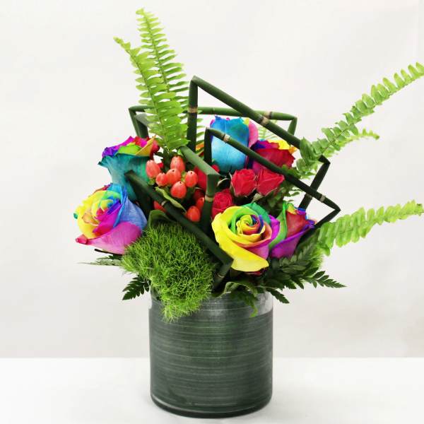Rainbow roses with red spray roses and ferns in a green vase with geometric bamboo accents