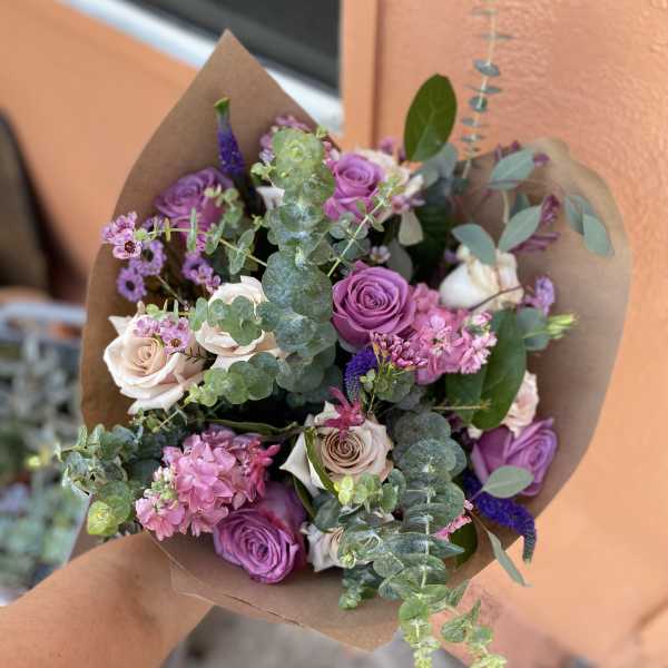Handheld bouquet of purple and blush roses with eucalyptus in brown paper