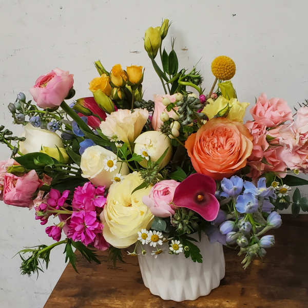 Low arrangement of mixed pastel roses, ranunculus, calla lily, and other blooms in a white vase.