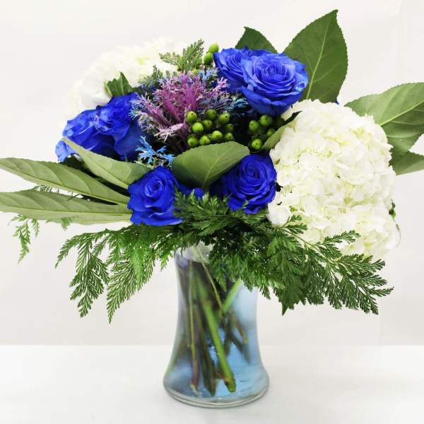 Blue roses and white hydrangeas with greenery in a clear glass vase with blue-tinted water