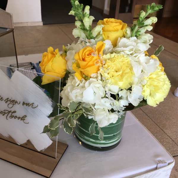Yellow and white flower arrangement with roses and snapdragons in a green vase beside an acrylic guestbook sign