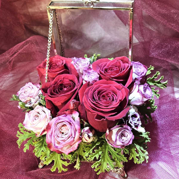 Compact arrangement of red and lavender roses in a gold metal and glass box on a burgundy fabric background