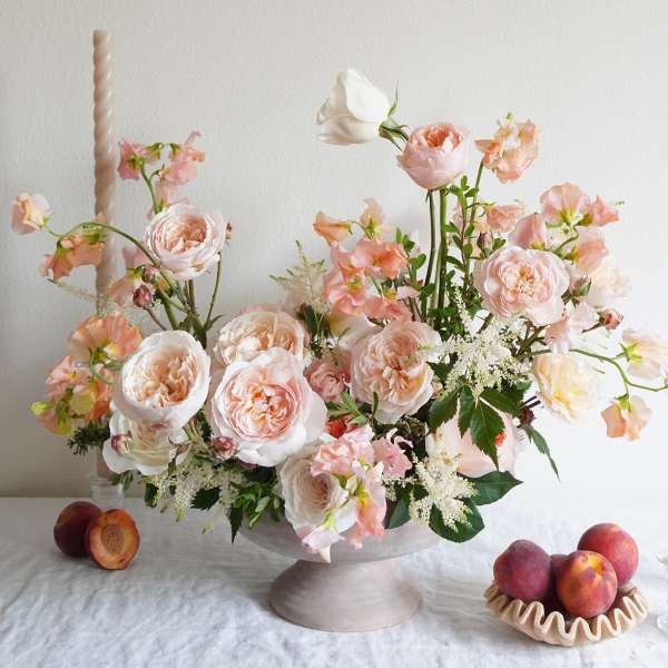 Blush roses and peach flowers arranged in a pedestal bowl with peaches nearby.