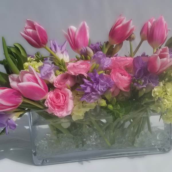 Low pink tulip, hydrangea, ranunculus, and purple freesia arrangement in a rectangular glass vase