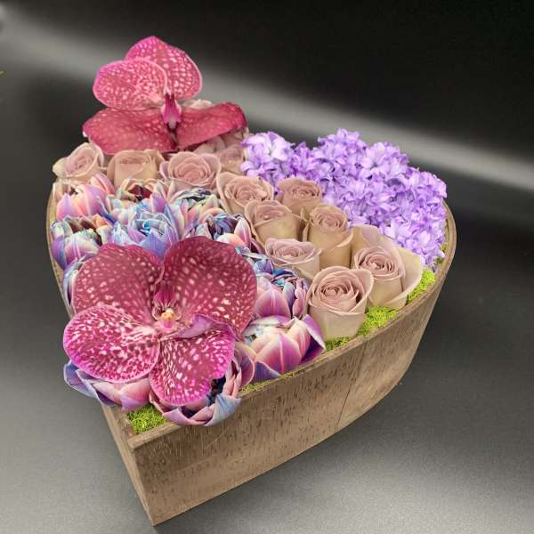 Heart-shaped flower arrangement with pink orchids, roses, and purple blooms in a wooden box
