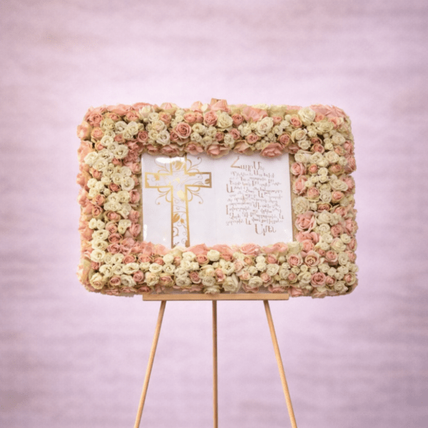 Rectangular frame of blush and cream roses around a printed cross panel on an easel.