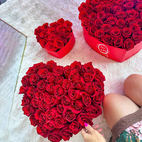 Heart-shaped red rose arrangements in red boxes on a soft white surface with a hand touching one box