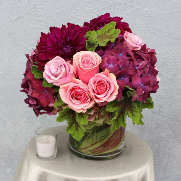 Pink roses and burgundy flowers in a glass vase with a candle beside it