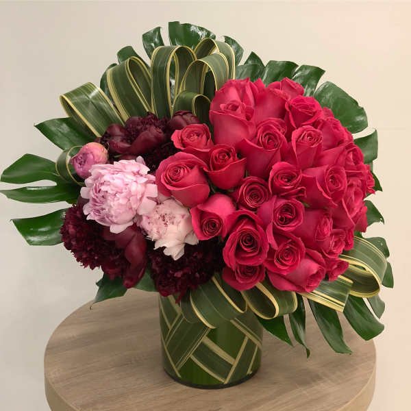 Hot pink roses with burgundy and blush blooms in a leaf-wrapped vase with looped striped foliage accents.