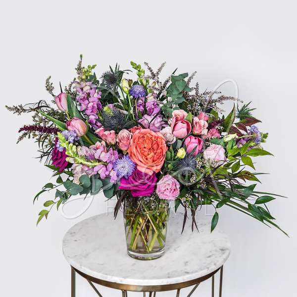 Lush arrangement of pink, purple, and peach flowers in a clear glass vase on a small round table.