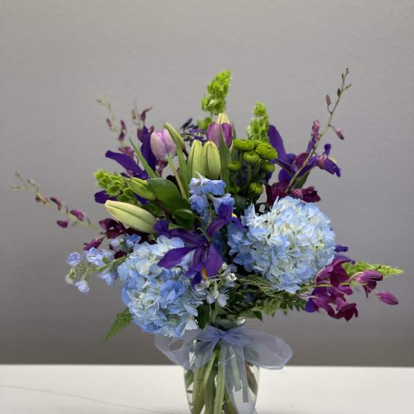 Mixed bouquet of blue hydrangeas, purple lilies, and green accents in a glass vase