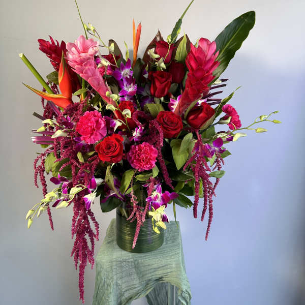 Tall tropical arrangement with red roses, hot pink blooms, and purple orchids in a green cylinder vase