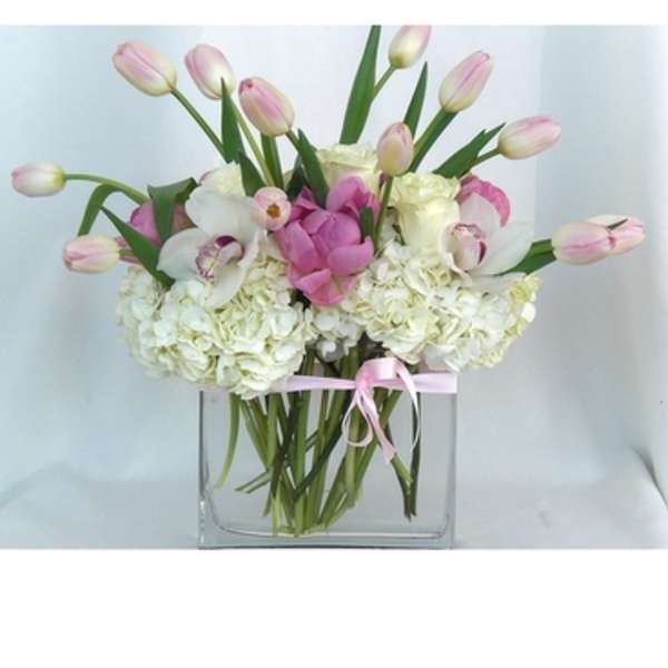Pink tulips and white flowers arranged in a clear square vase with a pink ribbon