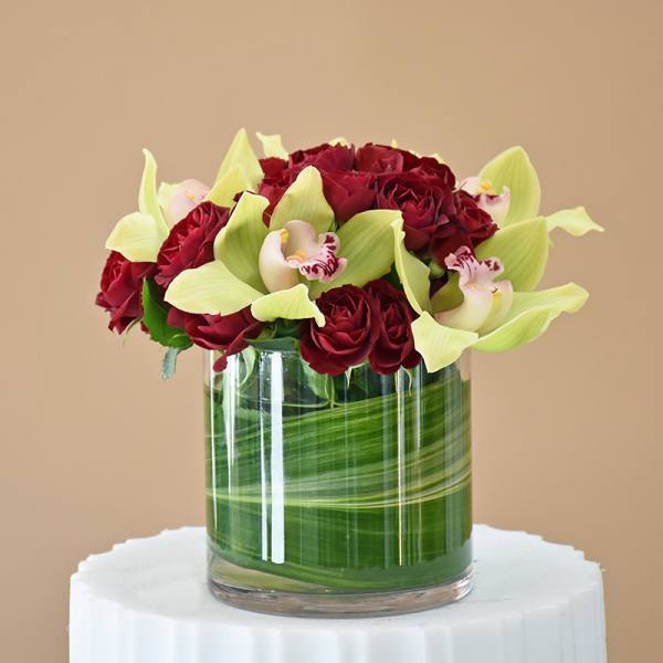 Red roses and pale green orchids arranged in a glass cylinder vase.