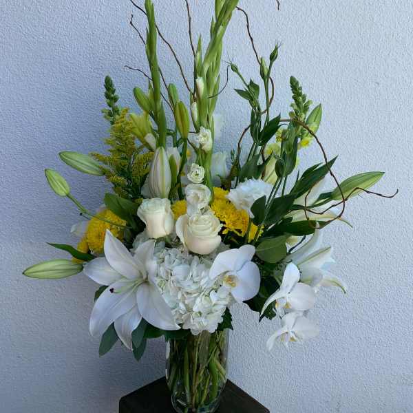 Tall white floral arrangement in a clear glass vase with lilies, roses, and orchids