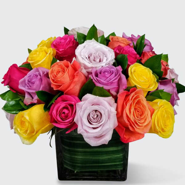 Compact arrangement of multicolored roses in a leaf-lined square glass vase