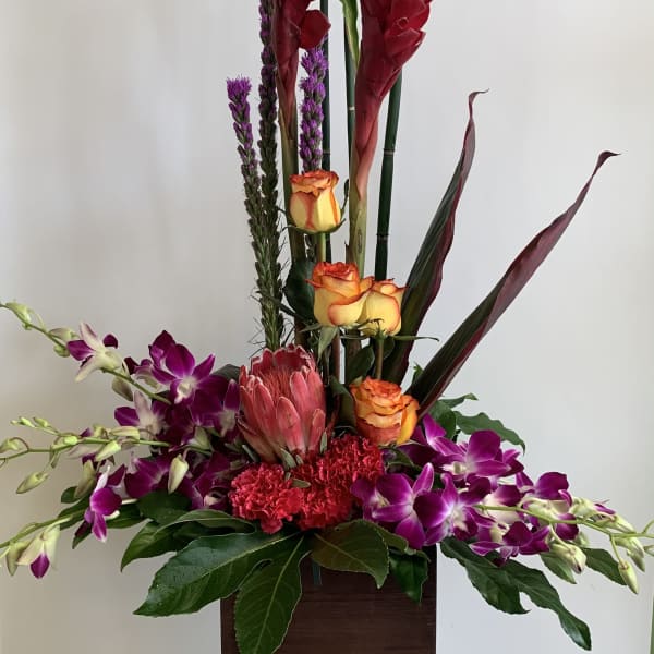 Tall tropical arrangement with red blooms, orange roses, and purple orchids in a wooden container