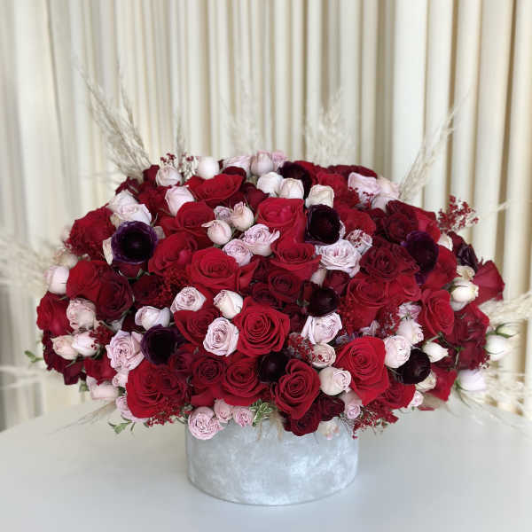 Low round arrangement of red, pink, and burgundy roses in a gray cylinder vase with pampas accents