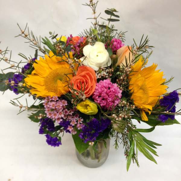 Mixed bouquet with sunflowers, roses, and purple filler in a glass vase