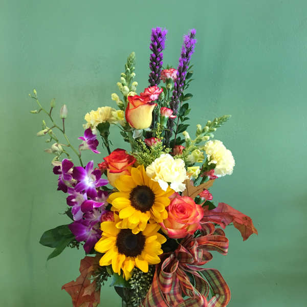Mixed bouquet with sunflowers, roses, orchids, and purple spikes in a glass vase