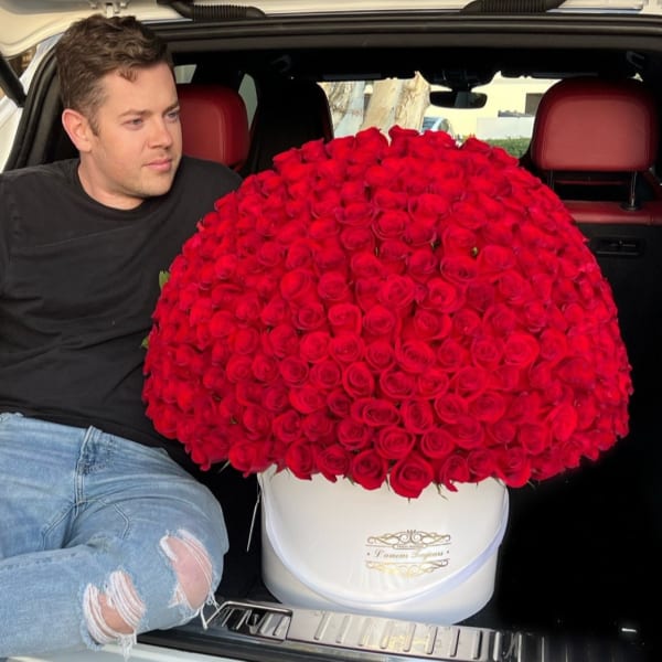 Large dome of red roses in a white hatbox in a car trunk beside a seated man