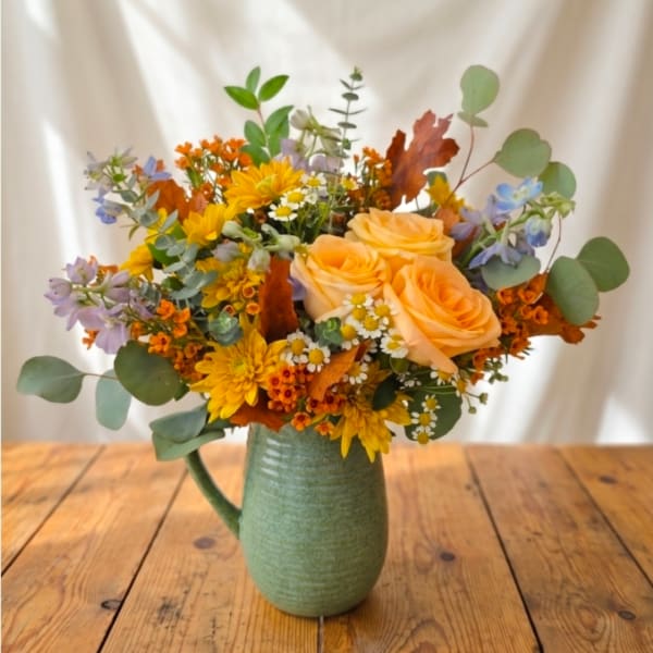 Bouquet of peach roses, yellow daisies, and orange flowers in a green pitcher vase