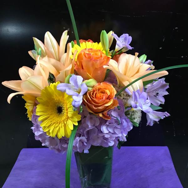 Mixed bouquet of orange, yellow, and lavender flowers in a glass vase